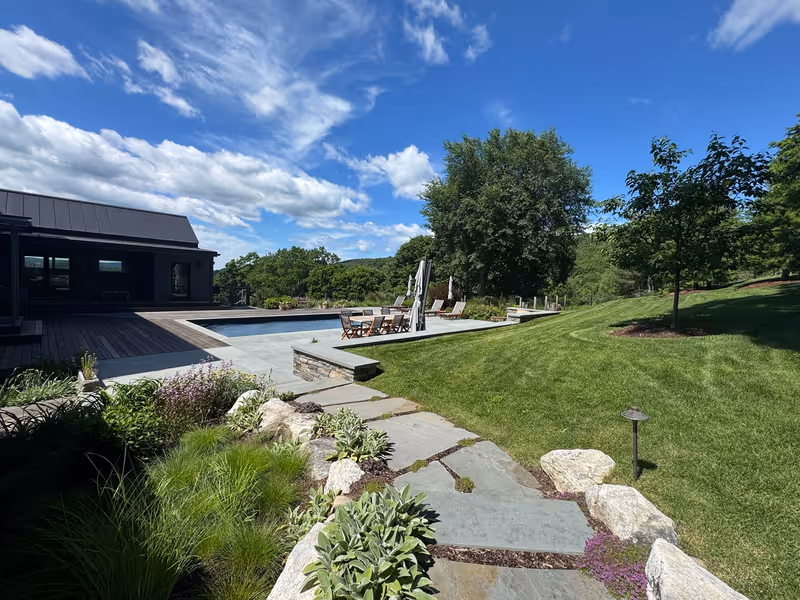 Stone path beside flowering borders, lawn and a swimming pool.