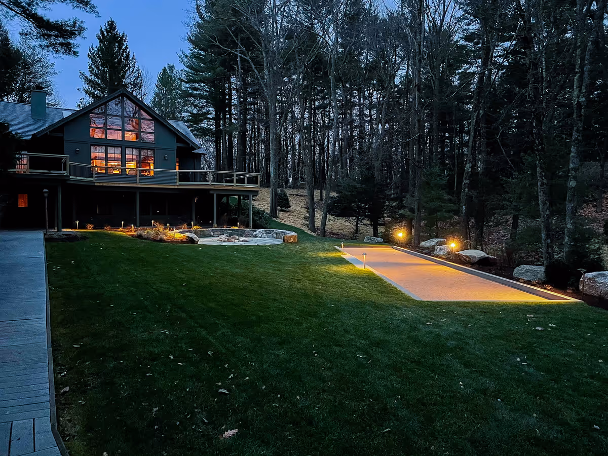 Illuminated bocce court beside lawn and a stone firepit area at dusk.