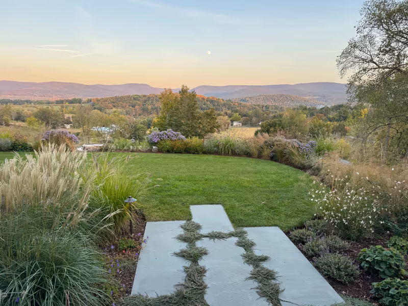 Stone path and planted borders opening onto a lawn and distant hills at sunset.