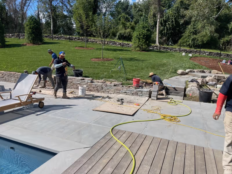 Crew installing stone paving and steps beside a residential swimming pool.