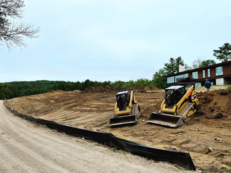 Two tracked loaders on a broad excavated slope beside a residence, with sediment fencing below.