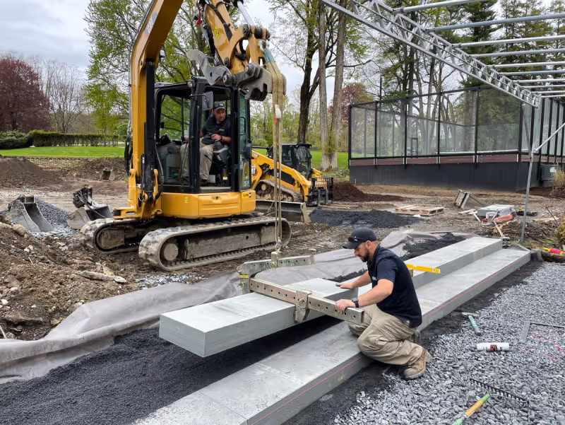 Mason positioning a large stone tread with an excavator beside a pool terrace.