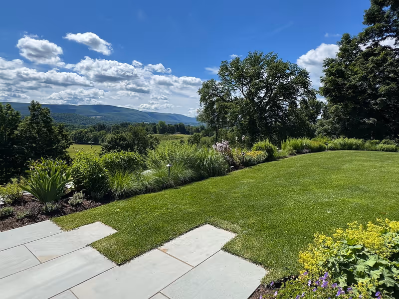 Perennial border beside a mown lawn and stone paving overlooking rural hills.
