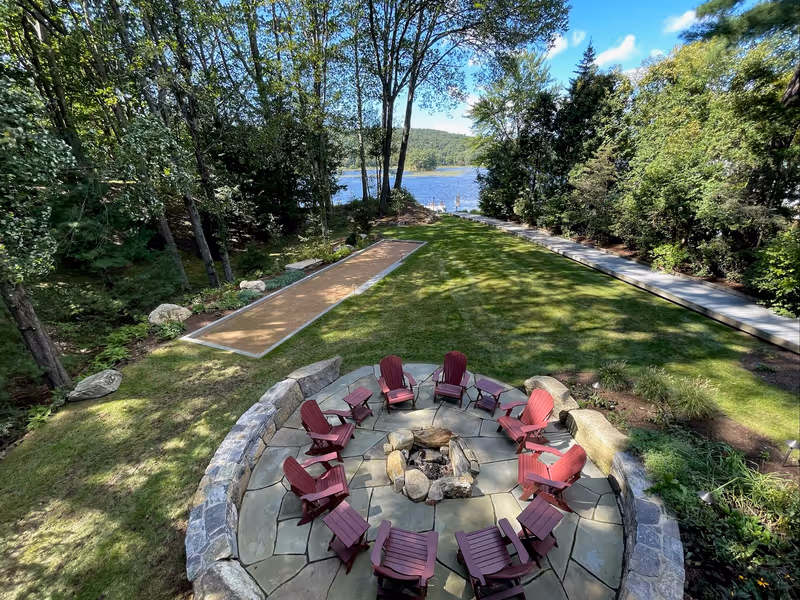 Stone fire pit and circular seating terrace on wooded lakeside grounds.