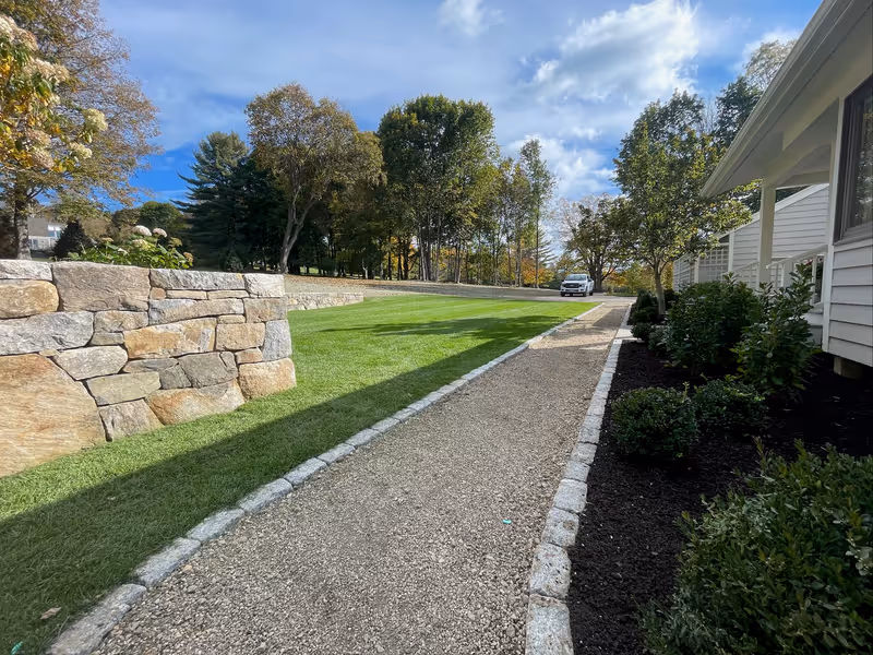 Stone walls and sod lawn beside an edged gravel walk and planting along a white house.