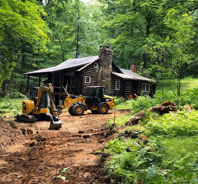 Excavator and loader working beside a timber house in a wooded setting.
