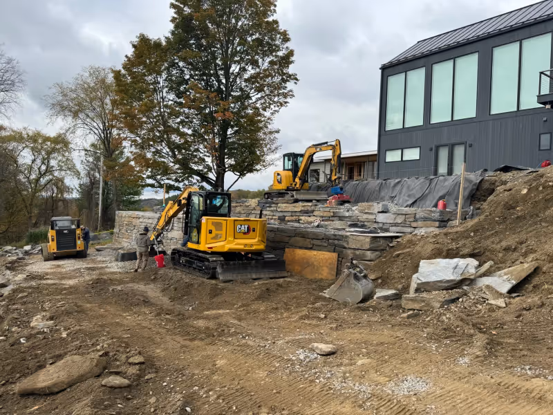 Tiered Goshen stone walls under construction with excavator and stone on site.